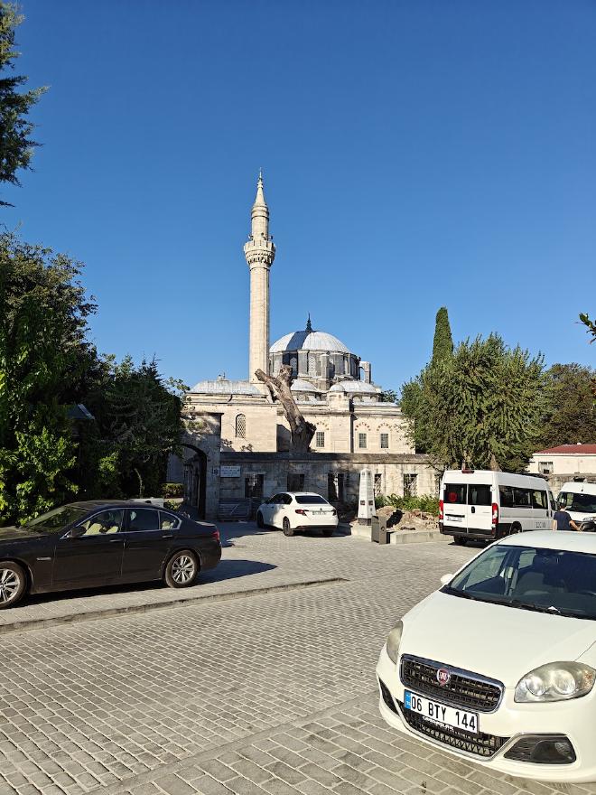 A small neighborhood mosque with a single minaret on a quiet Istanbul street, with parked cars in the foreground.