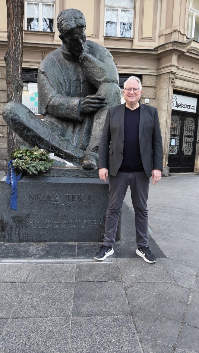 Kristian Stoffregen standing beside the seated bronze statue of Nikola Tesla in Zagreb, a wreath resting at the base of the monument.