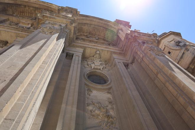 The Granada Cathedral facade seen from below, with carved relief panels and an oculus window in sharp afternoon light.