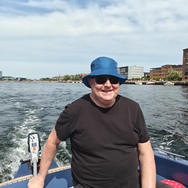 Kristian Stoffregen at the helm of a small boat in Copenhagen harbor on a sunny day, wearing a blue bucket hat and sunglasses