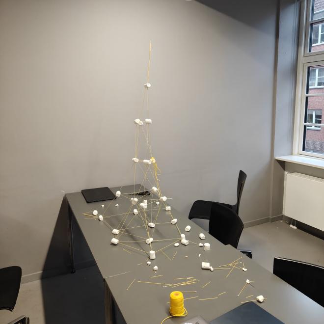 A finished tall spaghetti-and-marshmallow tower standing on a classroom desk by a window